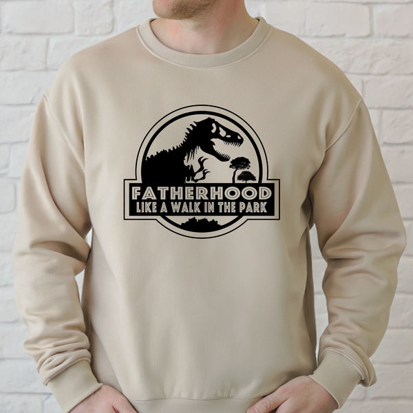 A man wearing a beige sweater with a graphic design featuring a dinosaur and the text "FATHERHOOD LIKE A WALK IN THE PARK".