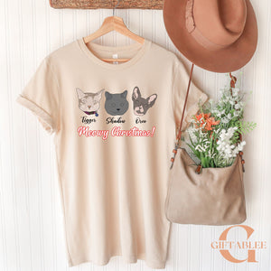 A beige t-shirt with a graphic design of three cat faces and the text "Meowy Christmas!" hanging on a wooden hanger, along with a brown hat and a beige purse.