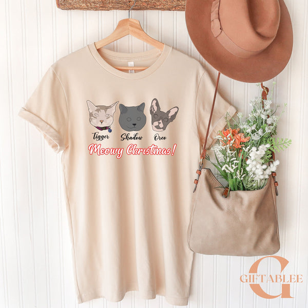 A beige t-shirt with a graphic design of three cat faces and the text "Meowy Christmas!" hanging on a wooden hanger, along with a brown hat and a beige purse.