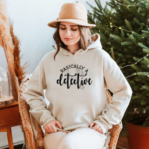 A woman wearing a beige hat and a white hoodie with the text "basically a detective" is sitting in a wicker chair in front of a Christmas tree.