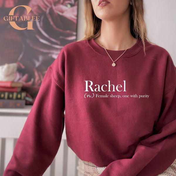 A woman wearing a maroon sweatshirt with the word "Rachel" printed on it, standing in front of a bookshelf.