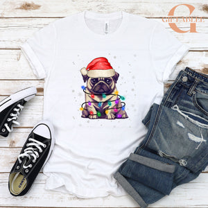 A white t-shirt with a graphic of a pug wearing a Santa hat, surrounded by Christmas lights, placed on a wooden surface alongside a pair of jeans and sneakers.