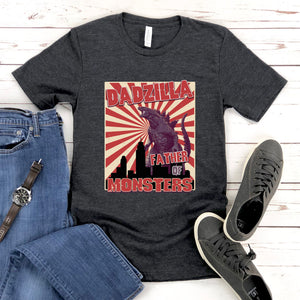 a gray t-shirt with a graphic design of a large purple monster, along with the text "DADZILLA" and "FATHER OF MONSTERS" printed on it. The t-shirt is laid out on a wooden surface, accompanied by a pair of jeans and a pair of sneakers.