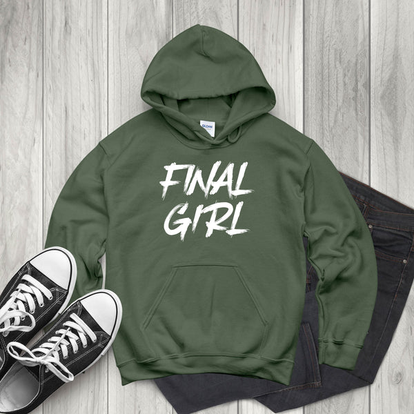 A green hoodie with the text "FINAL GIRL" printed on it, along with a pair of black and white sneakers and a pair of dark jeans, all laid out on a wooden surface.