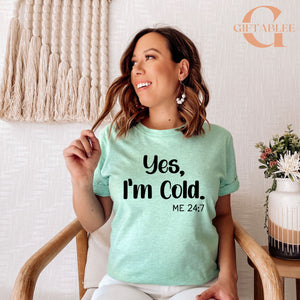 A woman wearing a light green t-shirt with the text "Yes, I'm Cold" and "ME 24.7" printed on it, smiling and looking to the side.