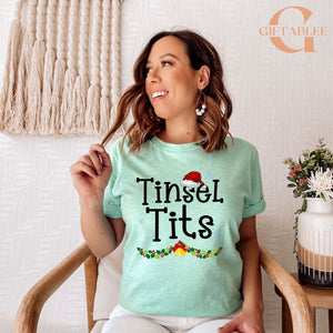 A woman wearing a light green t-shirt with the text "Tinsel Tits" is sitting in a chair, smiling and holding her hair.