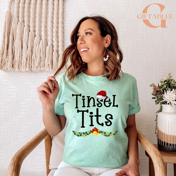 A woman wearing a light green t-shirt with the text "Tinsel Tits" is sitting in a chair, smiling and holding her hair.