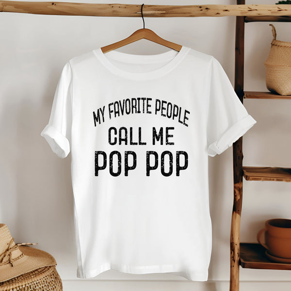 A white t-shirt with the text "MY FAVORITE PEOPLE CALL ME POP POP" printed on it, hanging on a wooden hanger.