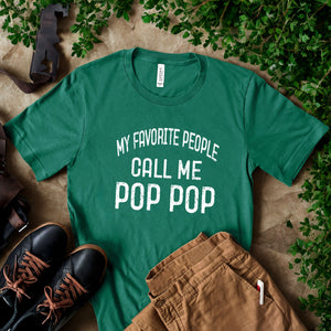 A green t-shirt with the text "MY FAVORITE PEOPLE CALL ME POP POP" printed on it, along with a pair of brown pants and black shoes, all laid out on a surface with some greenery.