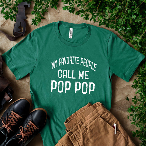 A green t-shirt with the text "MY FAVORITE PEOPLE CALL ME POP POP" printed on it, along with a pair of brown pants and black shoes, all laid out on a surface with some greenery.