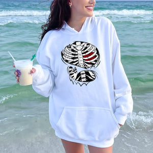 A woman wearing a white hoodie with a graphic design stands on a beach, holding a drink in her hand.
