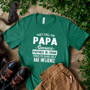 A green t-shirt with white text that reads "They call me PAPA because Partner in Crime makes me sound like a bad influence" is displayed on a table, along with a pair of black shoes and some greenery.