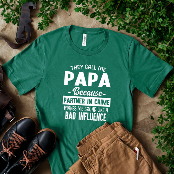 A green t-shirt with white text that reads "They call me PAPA because Partner in Crime makes me sound like a bad influence" is displayed on a table, along with a pair of black shoes and some greenery.