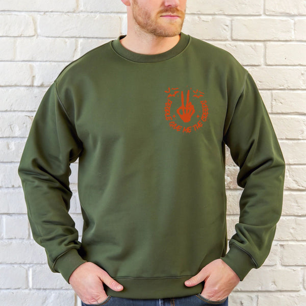 A man wearing a green sweatshirt with an orange graphic on it stands in front of a white brick wall.