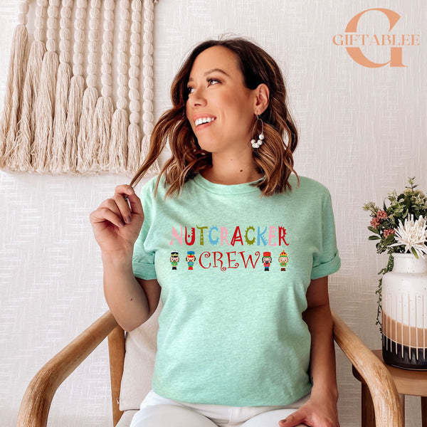A woman wearing a mint green t-shirt with the text "NUTCRACKER CREW" is sitting in a chair, smiling and holding her hair.