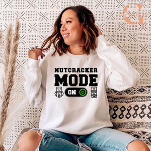 A woman wearing a white sweatshirt with the text "NUTCRACKER MODE ON" printed on it, sitting on a patterned couch.