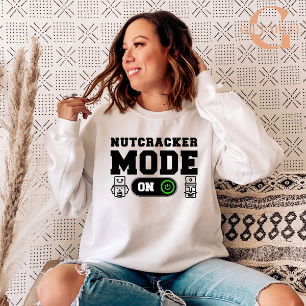 A woman wearing a white sweatshirt with the text "NUTCRACKER MODE ON" printed on it, sitting on a patterned couch.