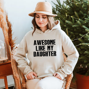 A woman wearing a hat and a white hoodie with the text "AWESOME LIKE MY DAUGHTER" is sitting in a chair in front of a Christmas tree.