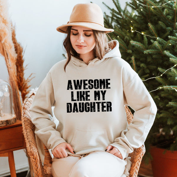 A woman wearing a hat and a white hoodie with the text "AWESOME LIKE MY DAUGHTER" is sitting in a chair in front of a Christmas tree.