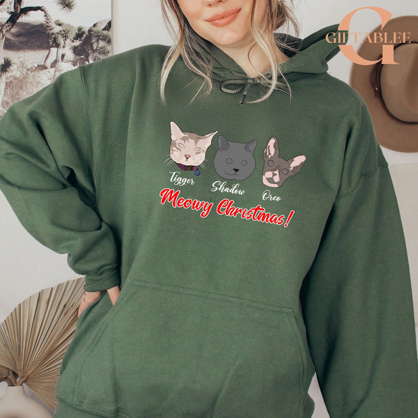 A woman wearing a green hoodie with a graphic design of three cat faces and the text "Meowy Christmas!" printed on it.