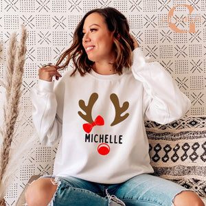 A woman wearing a white sweater with a graphic of a reindeer and the name "Michele" printed on it, smiling and looking to the side.