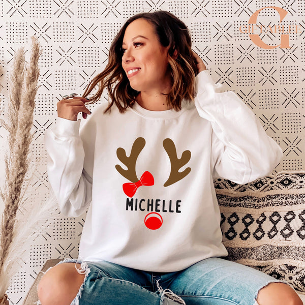 A woman wearing a white sweater with a graphic of a reindeer and the name "Michele" printed on it, smiling and looking to the side.