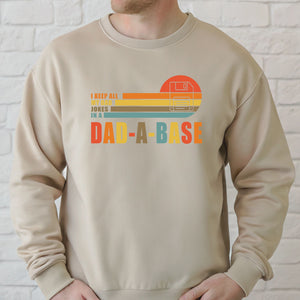 A man wearing a beige sweater with a graphic design that says "I KEEP ALL MY JOKES IN A DAD-A-BASE" printed on it.