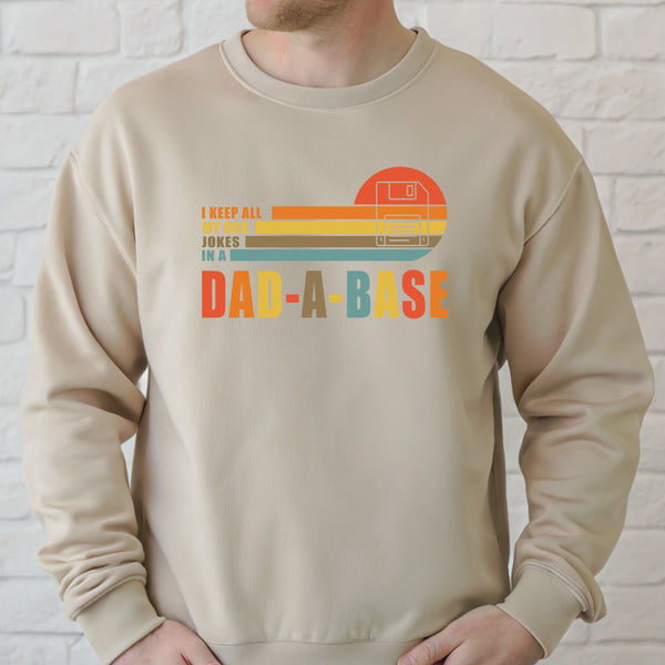 A man wearing a beige sweater with a graphic design that says "I KEEP ALL MY JOKES IN A DAD-A-BASE" printed on it.