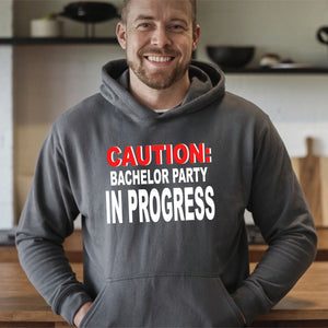 A man wearing a grey hoodie with the text "CAUTION: BACELOR PARTY IN PROGRESS" printed on it.