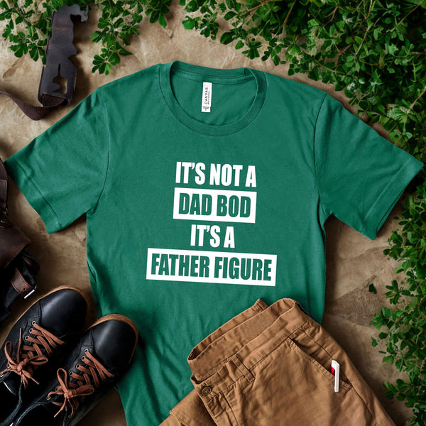 A green t-shirt with white text that reads "IT'S NOT A DAD BOD IT'S A FATHER FINE" is displayed on a surface with some clothing items and a pair of shoes.