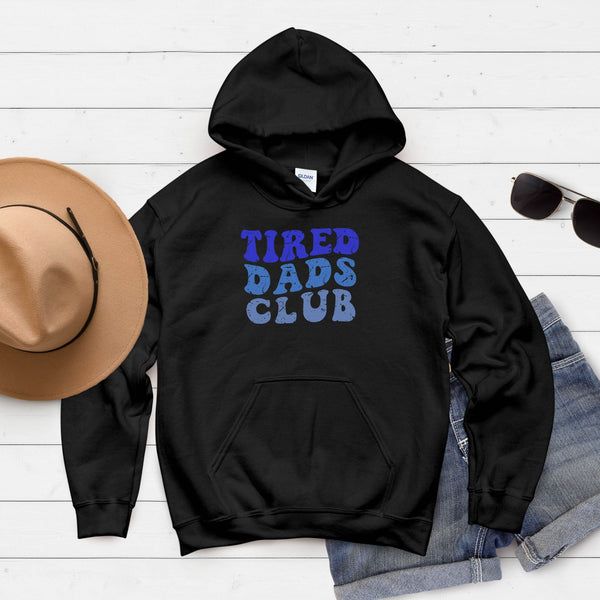A black hoodie with the text "TIRED DAD'S CLUB" printed on it, along with a pair of sunglasses and a hat, all laid out on a white wooden surface.