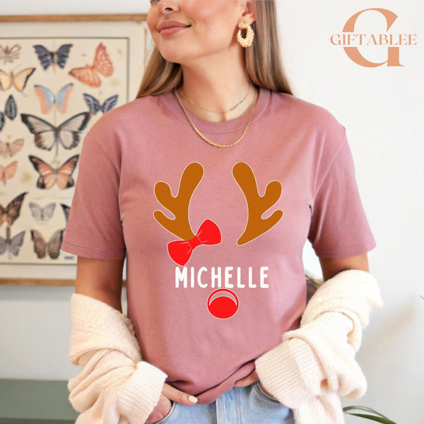 A woman wearing a pink t-shirt with a reindeer and the name "Michele" printed on it, standing in front of a butterfly-themed wall art.