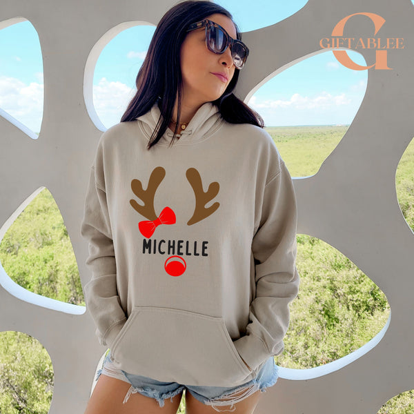 A woman wearing a grey hoodie with a reindeer design and the name "Michele" printed on it, standing in front of a window with a blue sky and trees visible outside.