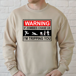 A person wearing a beige sweater with a graphic design that says "WARNING IF ZOMBIES CHASE US I'M TRIPPING YOU" along with various zombie-related icons.