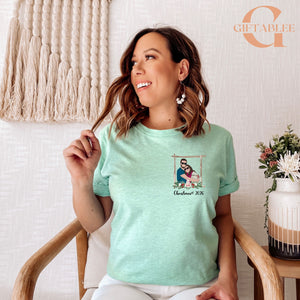 A woman wearing a mint green t-shirt with a Christmas-themed graphic is sitting in a chair, smiling and looking to the side.