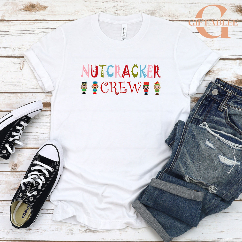 A white t-shirt with the text "NUTCRACKER CREW" printed on it, along with small nutcracker figures, is displayed on a wooden surface.