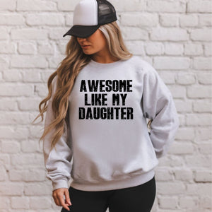 A woman wearing a gray sweatshirt with the text "AWESOME LIKE MY DAUGHTER" printed on it, standing in front of a white brick wall.