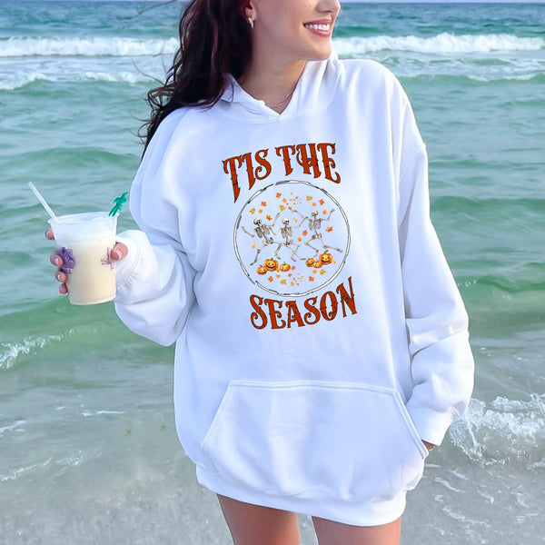 A woman wearing a white hoodie with a graphic design of dancing skeletons and the text "TIS THE SEASON" is standing on a beach, holding a drink.