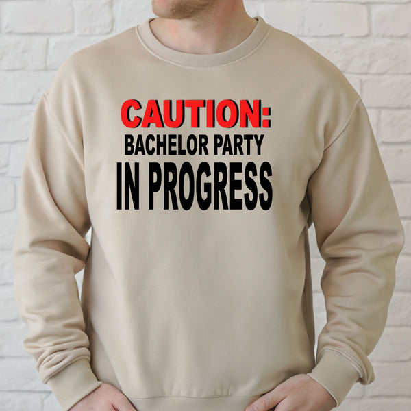 A man wearing a beige sweater with the text "CAUTION: BACCHELOR PARTY IN PROGRESS" printed on it.