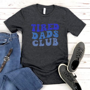A gray t-shirt with the text "TIRED DAD'S CLUB" printed on it, along with a pair of jeans and sneakers, all laid out on a wooden surface.