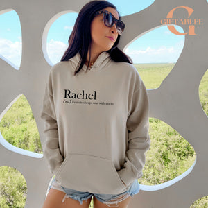 A woman wearing a grey hoodie with the word "Rachel" printed on it, standing in front of a window with a view of a field.