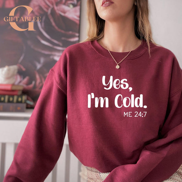 A woman wearing a maroon sweatshirt with the text "Yes, I'm Cold. ME 24:7" printed on it.