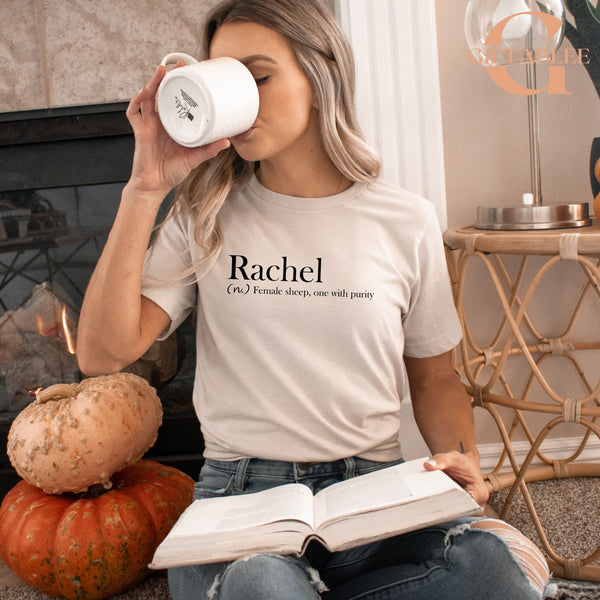 A woman is sitting on the floor, reading a book and drinking from a mug. She is wearing a white t-shirt with the word "Rachael" printed on it. There is a pumpkin and a lamp in the background, creating a cozy and autumnal atmosphere.