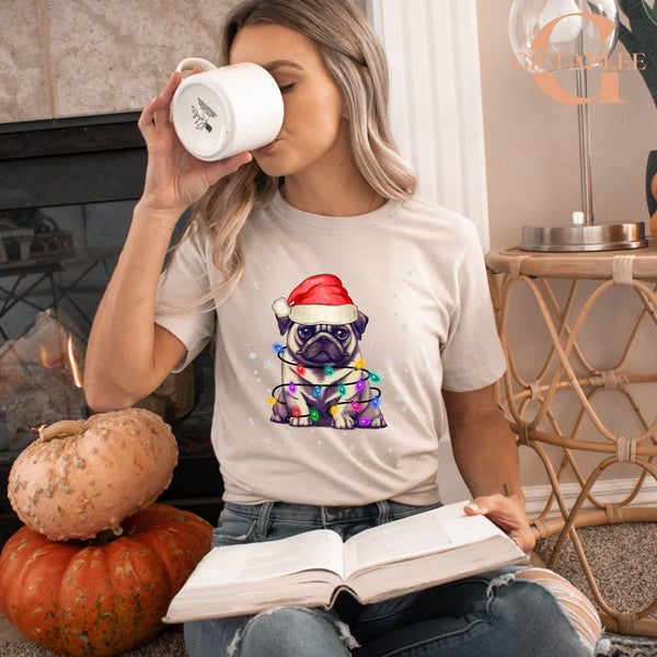 A woman is sitting on the floor, wearing a white t-shirt with a pug dog wearing a Santa hat and holding a book.