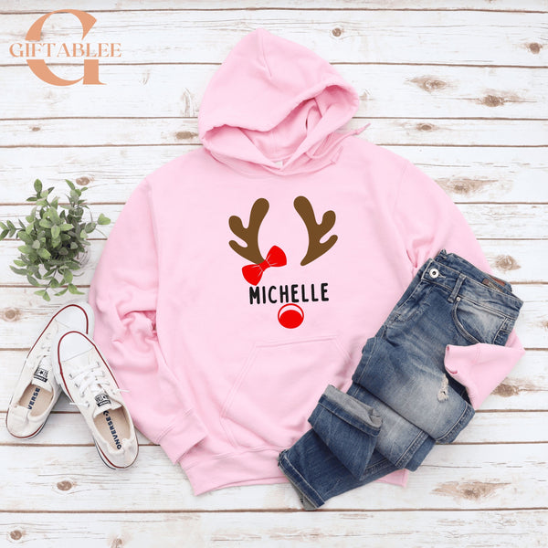 A pink hoodie with a graphic of a reindeer and the name "Michele" printed on it, along with a pair of jeans and sneakers, all laid out on a wooden surface.