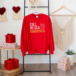 A red sweatshirt with the text "Call me old, fashioned" printed on it, hanging on a black hanger. The sweatshirt is displayed in a room with various decorations, including a macrame wall hanging, a red heart-shaped ornament, and a stack of wrapped presents.
