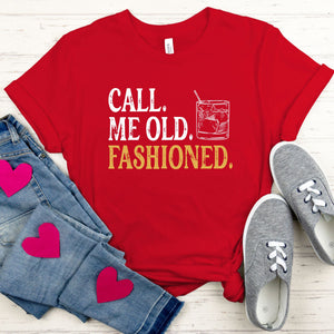 A red t-shirt with the text "CALL, ME OLD, FASHIONED." printed on it, along with a graphic of a glass of alcohol. The t-shirt is accompanied by a pair of jeans and gray sneakers, all laid out on a wooden surface.