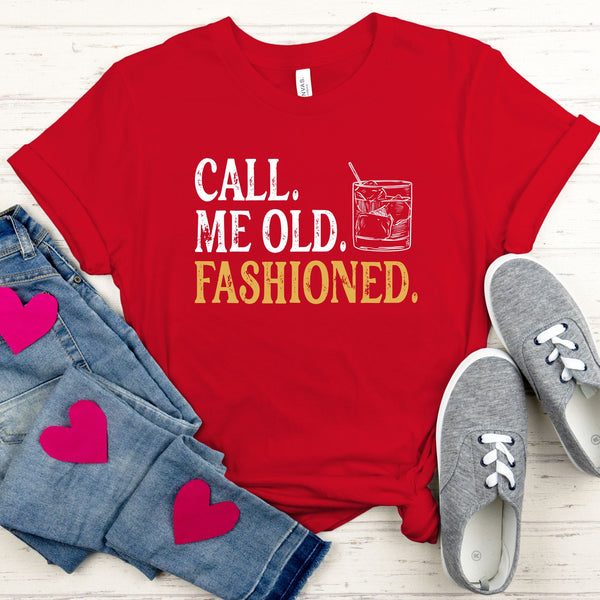 A red t-shirt with the text "CALL, ME OLD, FASHIONED." printed on it, along with a graphic of a glass of alcohol. The t-shirt is accompanied by a pair of jeans and gray sneakers, all laid out on a wooden surface.