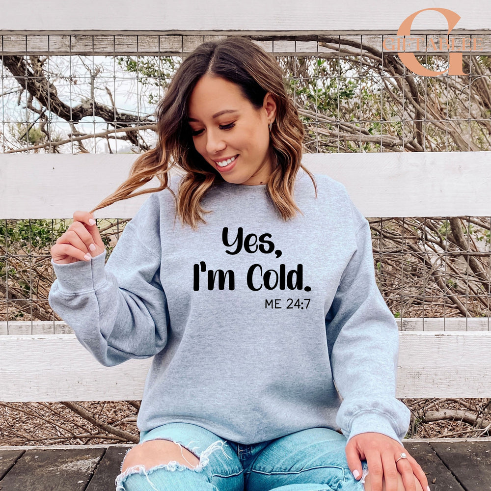 A woman wearing a gray sweatshirt with the text "Yes, I'm Cold. ME 24.7" is sitting on a bench in front of a fence.