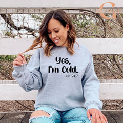A woman wearing a gray sweatshirt with the text "Yes, I'm Cold. ME 24.7" is sitting on a bench in front of a fence.